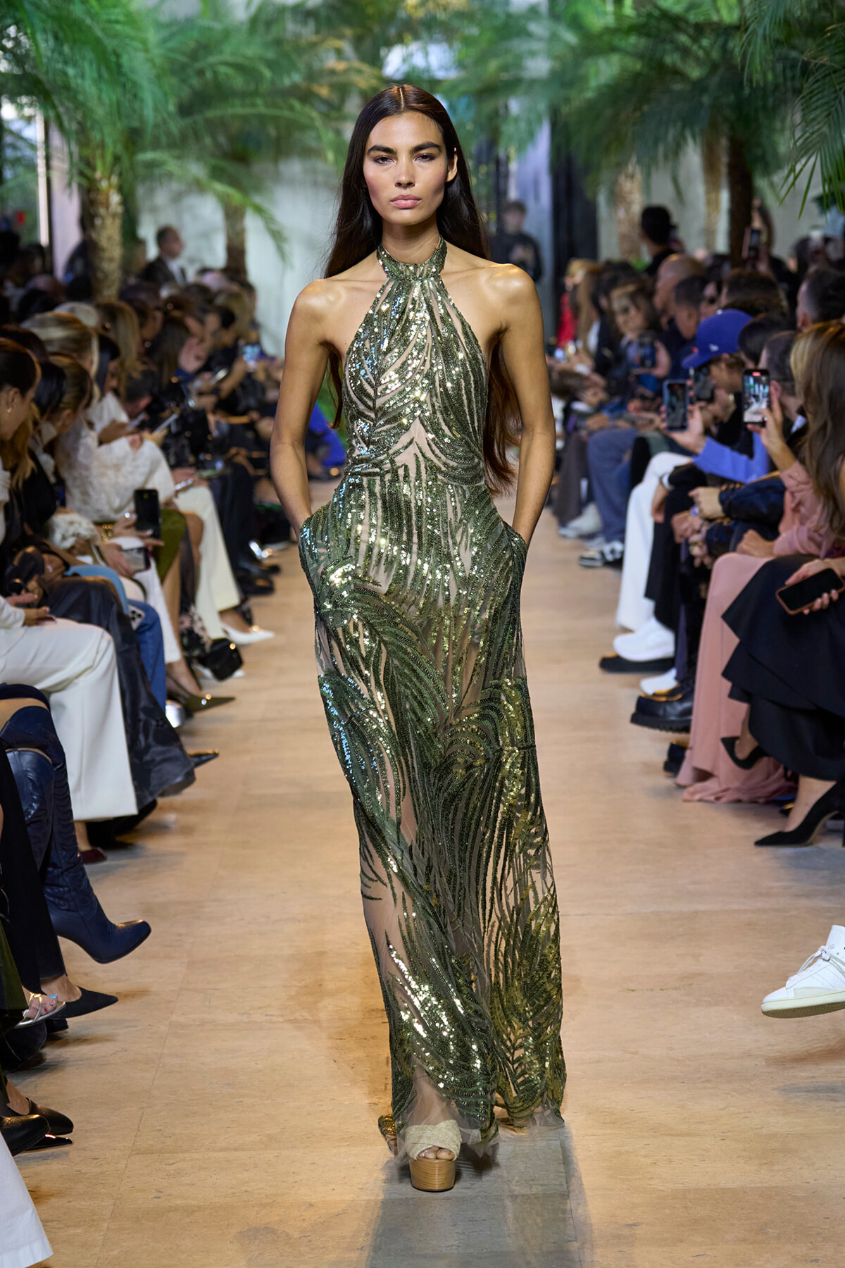 Model walks runway in a shimmering green sequined halter-neck gown with palm backdrop and audience on either side.