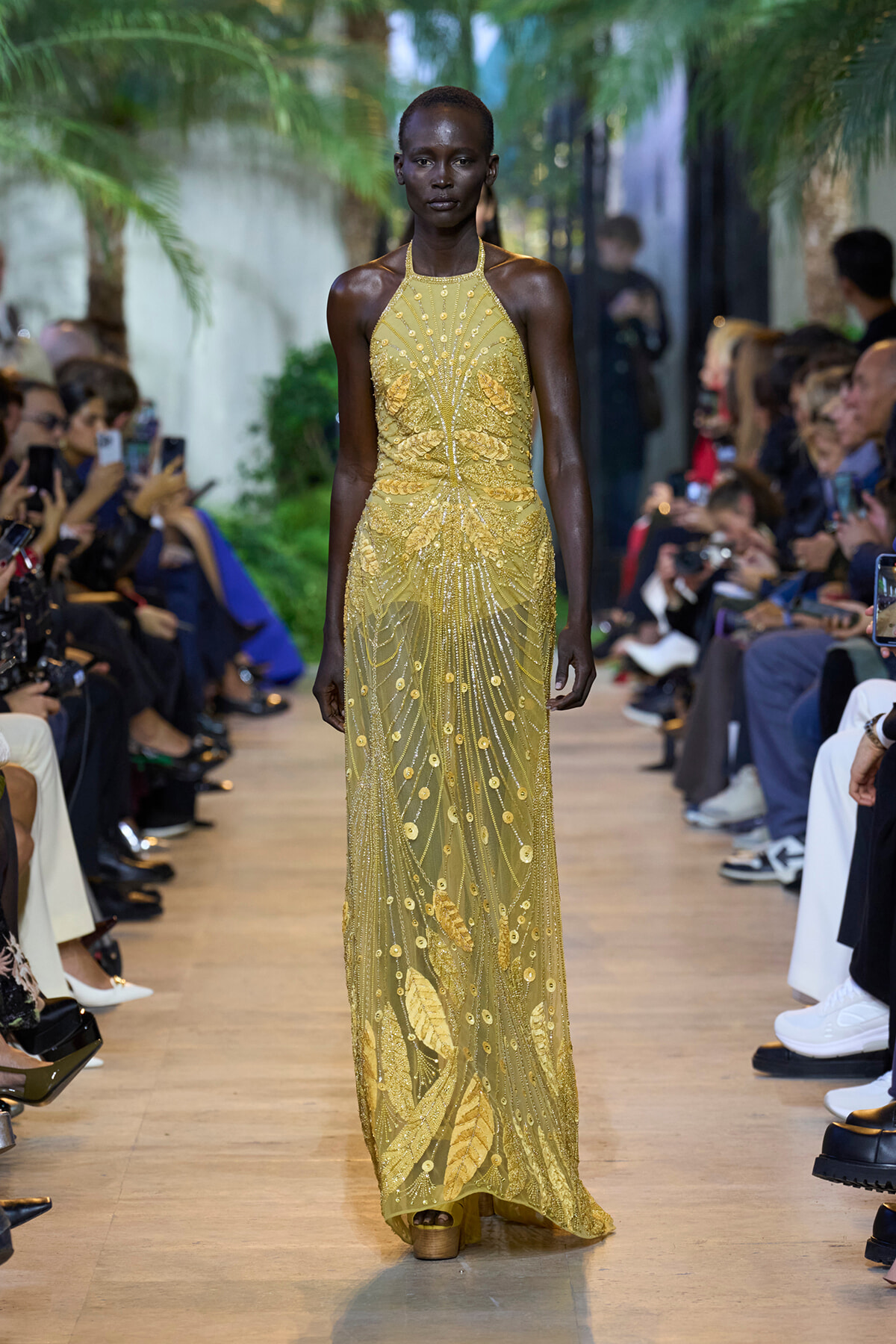 Model walks the runway in a gold beaded halter-neck gown with intricate leaf and line patterns, audience in the background.