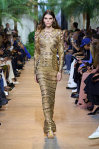 Model in a long, gold sequined gown walking a runway with spectators on both sides.