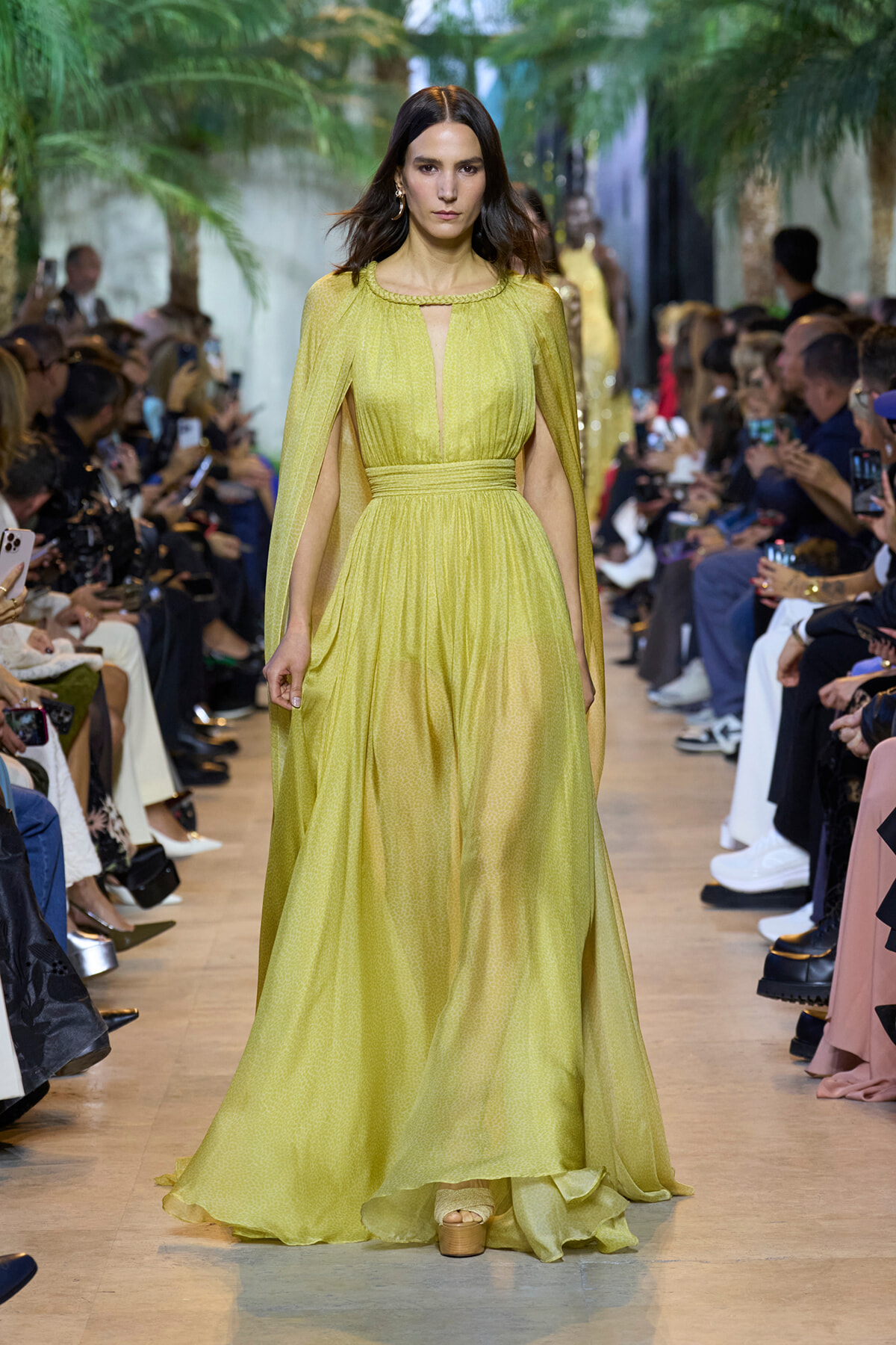 Model walks the runway in a lime-yellow cape gown with a gathered waist; audience on both sides taking photos.