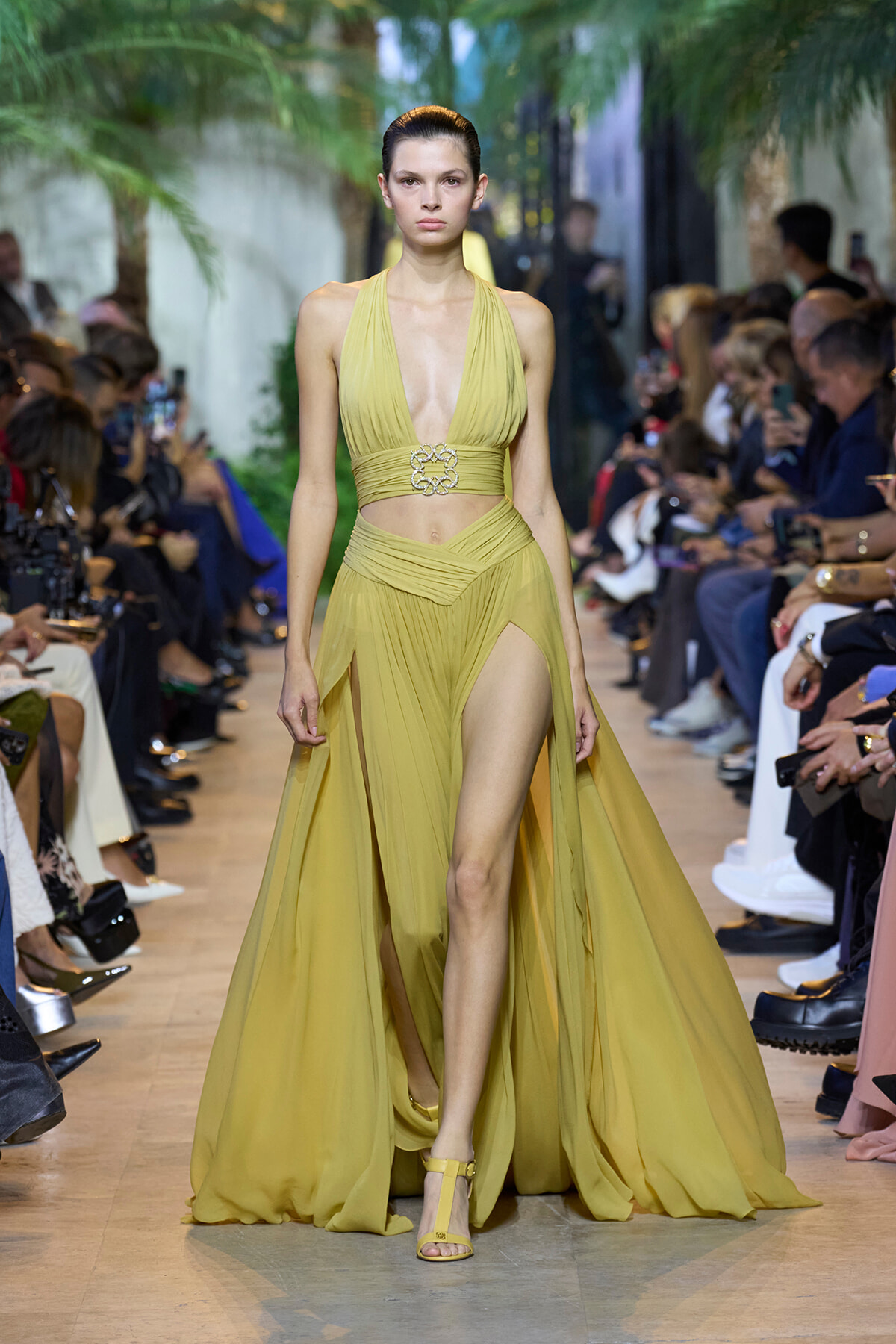 Model walking a runway in a yellow halter gown with a deep V neckline and thigh-high slit, surrounded by spectators.