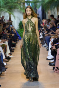 Model walking a runway in a shimmering green halter gown with a front cutout and gathered waist, audience on both sides watching.