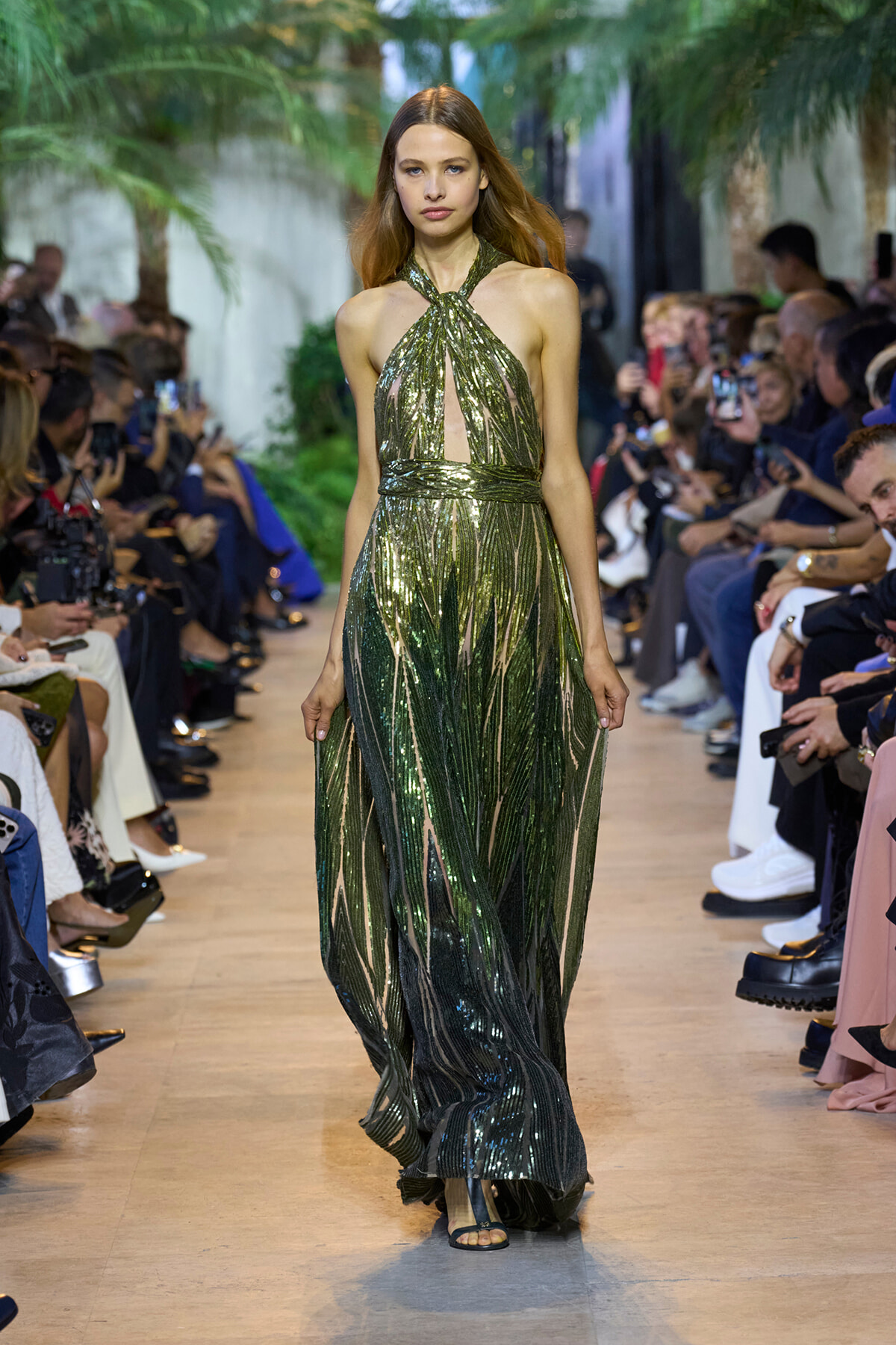 Model walking a runway in a shimmering green halter gown with a front cutout and gathered waist, audience on both sides watching.