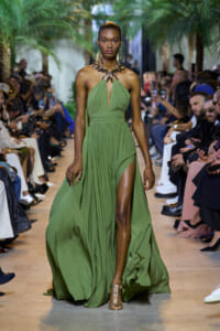 Model walks a green halter gown with deep V neckline and high slit down the left leg on a fashion runway, wearing a bold metallic necklace and gold sandals.