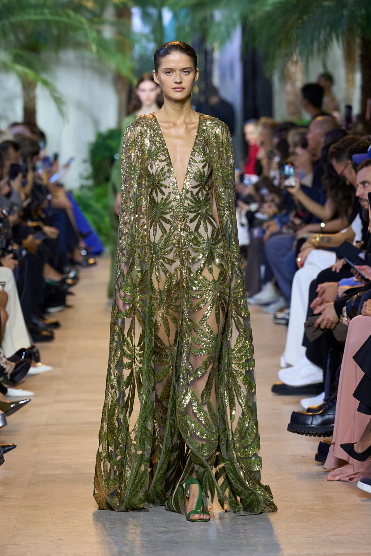 Model walks a runway in a sheer, emerald gown heavily embellished with leafy green sequins and a long flowing cape-like overlay, green sandals visible