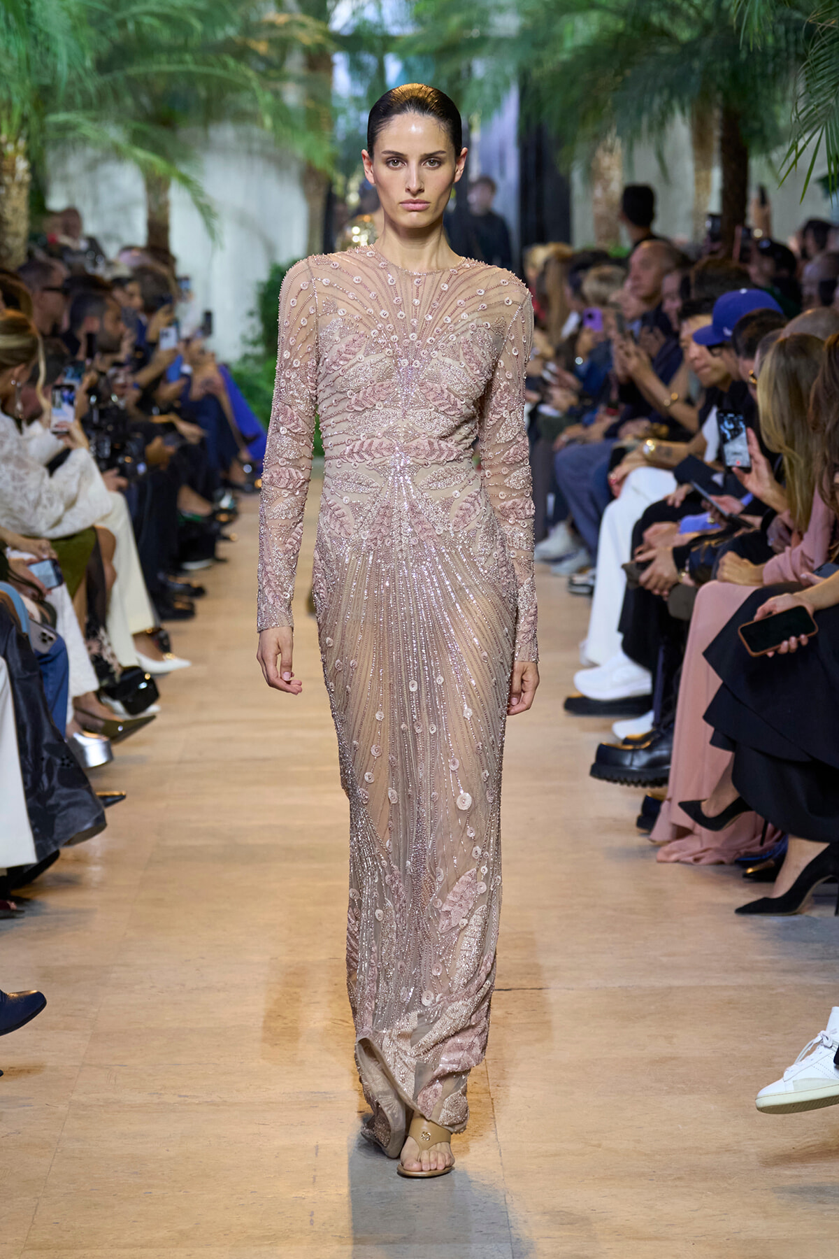 Model walking a runway in a long nude-pink beaded gown with intricate embroidery, surrounded by seated audience and photographers.