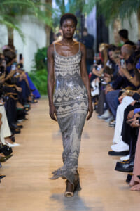 Model walks the runway in a silver-gray beaded gown with geometric patterns and fringe detailing.