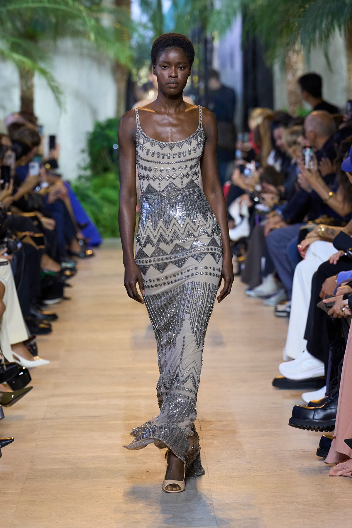 Model walks the runway in a silver-gray beaded gown with geometric patterns and fringe detailing.