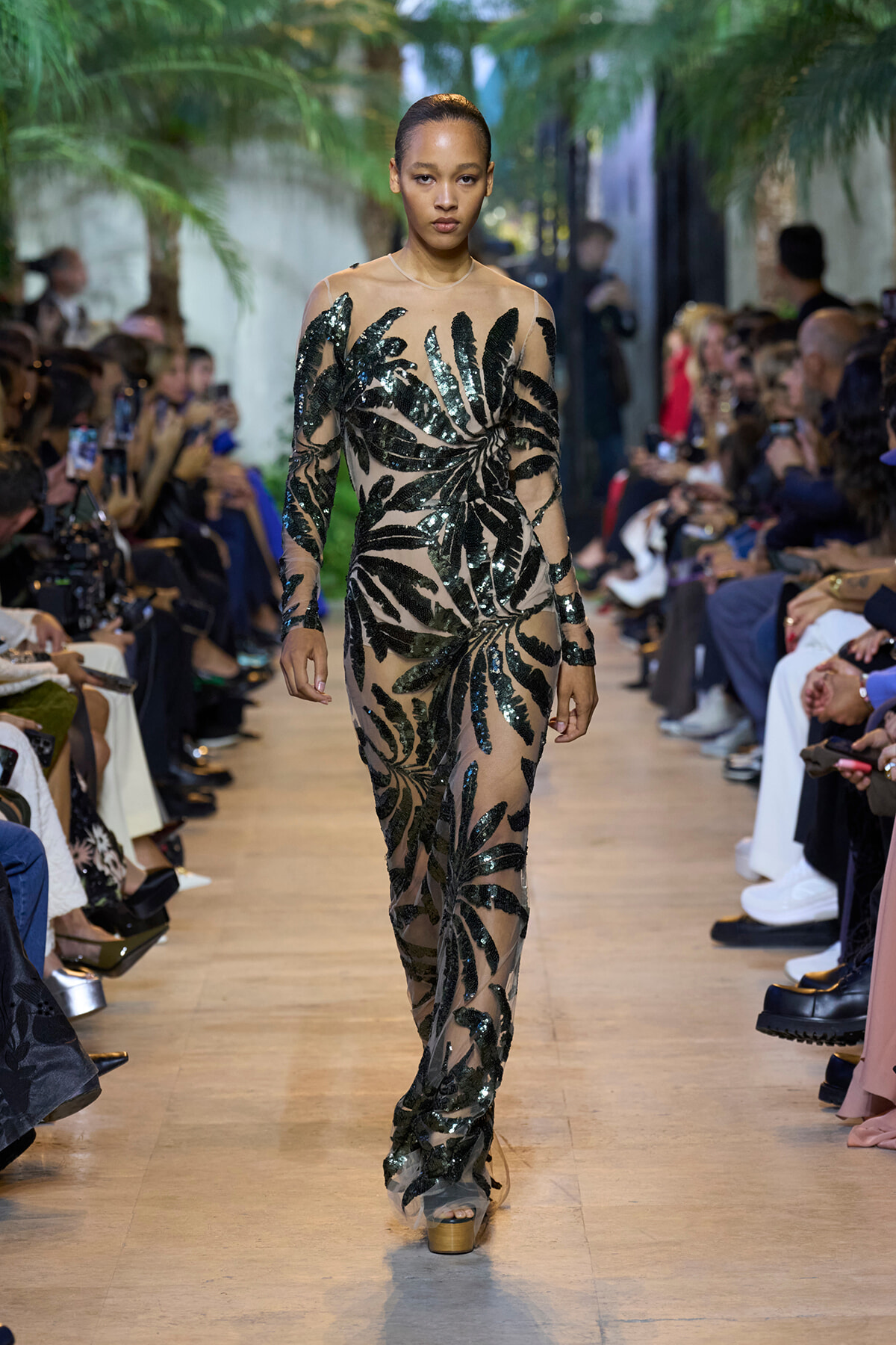 Model walks the runway in a sheer nude gown with dark sequined botanical leaf pattern, audience on both sides.