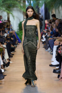 Model walks a runway in a strapless black lace gown with a dark green scarf draped around the neck, audience on both sides.
