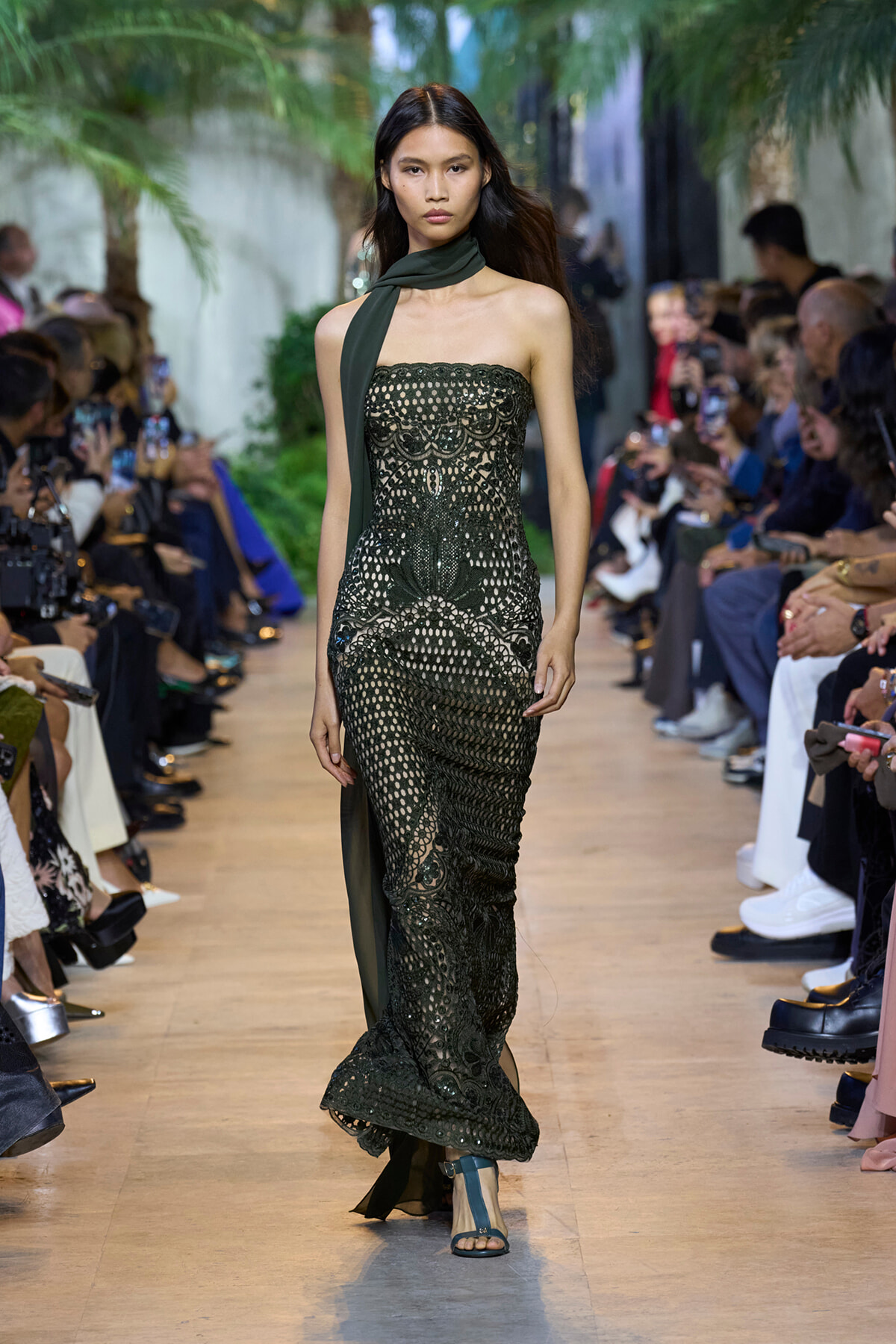 Model walks a runway in a strapless black lace gown with a dark green scarf draped around the neck, audience on both sides.