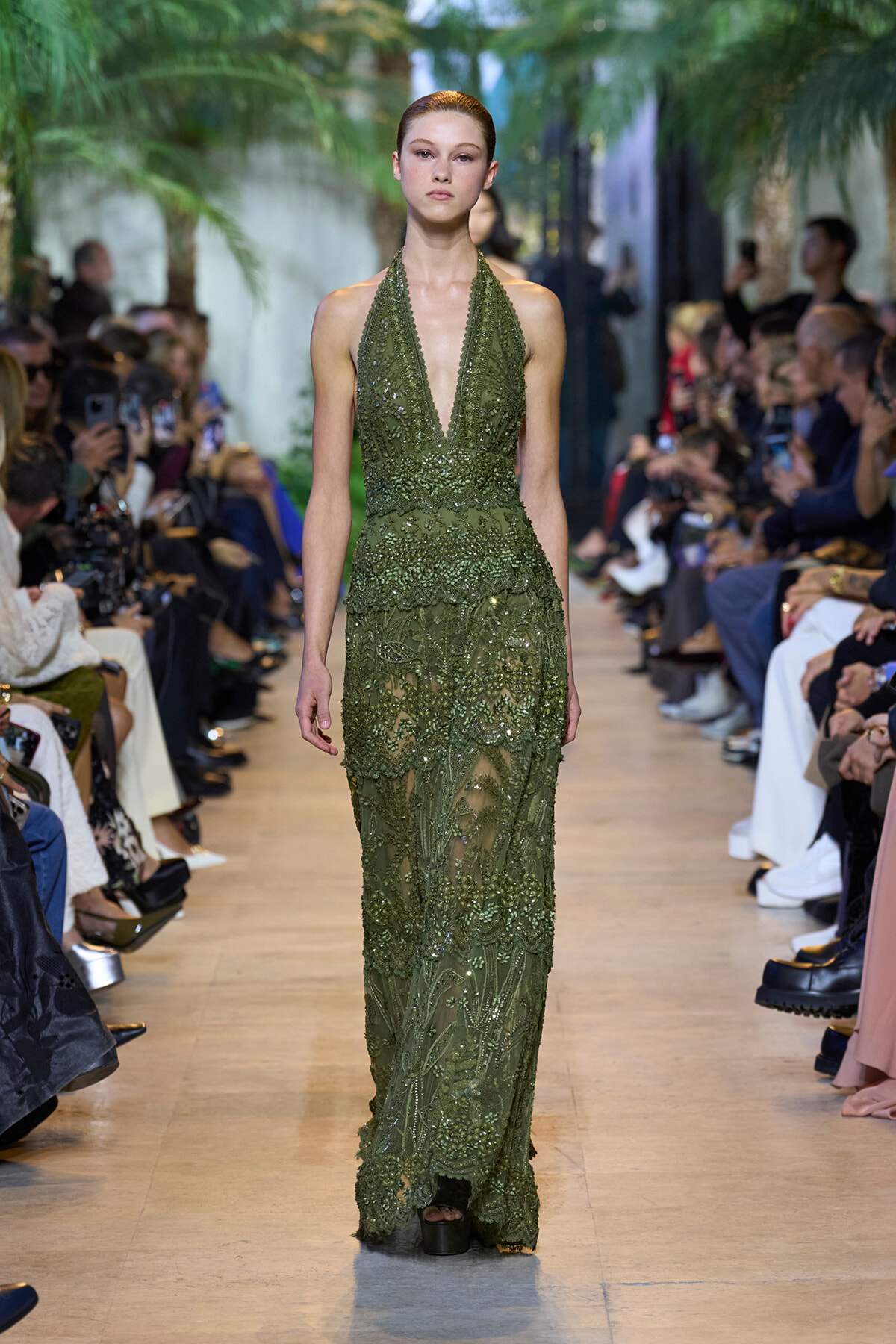 Model walks the runway in a floor-length olive green beaded lace gown with a plunging halter neckline.