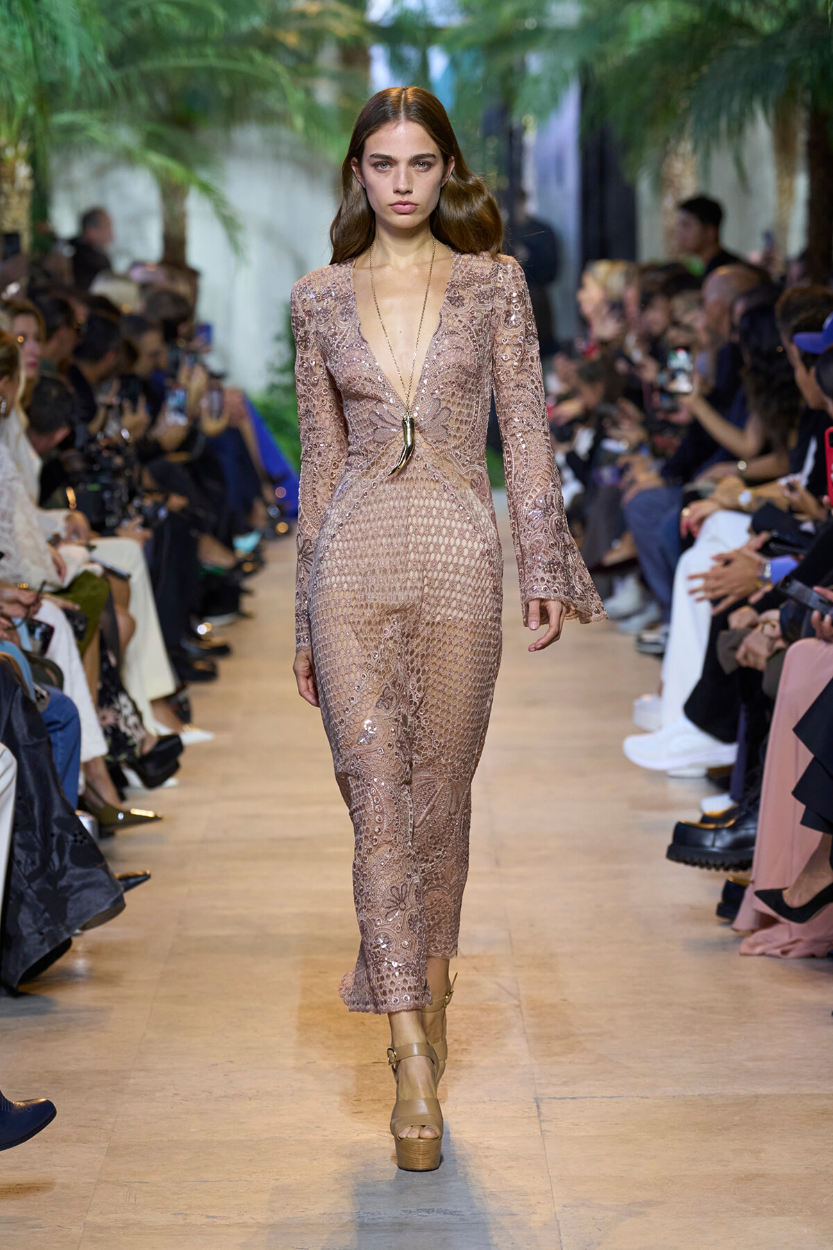 Model walks a runway in a dusty pink lace jumpsuit with a plunging V-neck, long bell sleeves, and platform tan sandals, while seated audience watches.