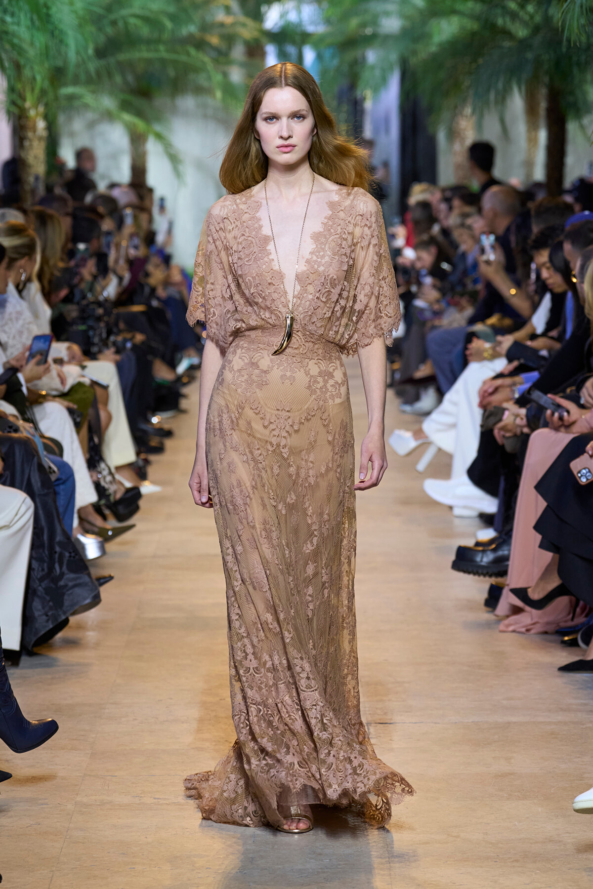 Model walks a beige lace gown down a fashion runway, wearing a long horn pendant necklace in a lush, green-lined venue.