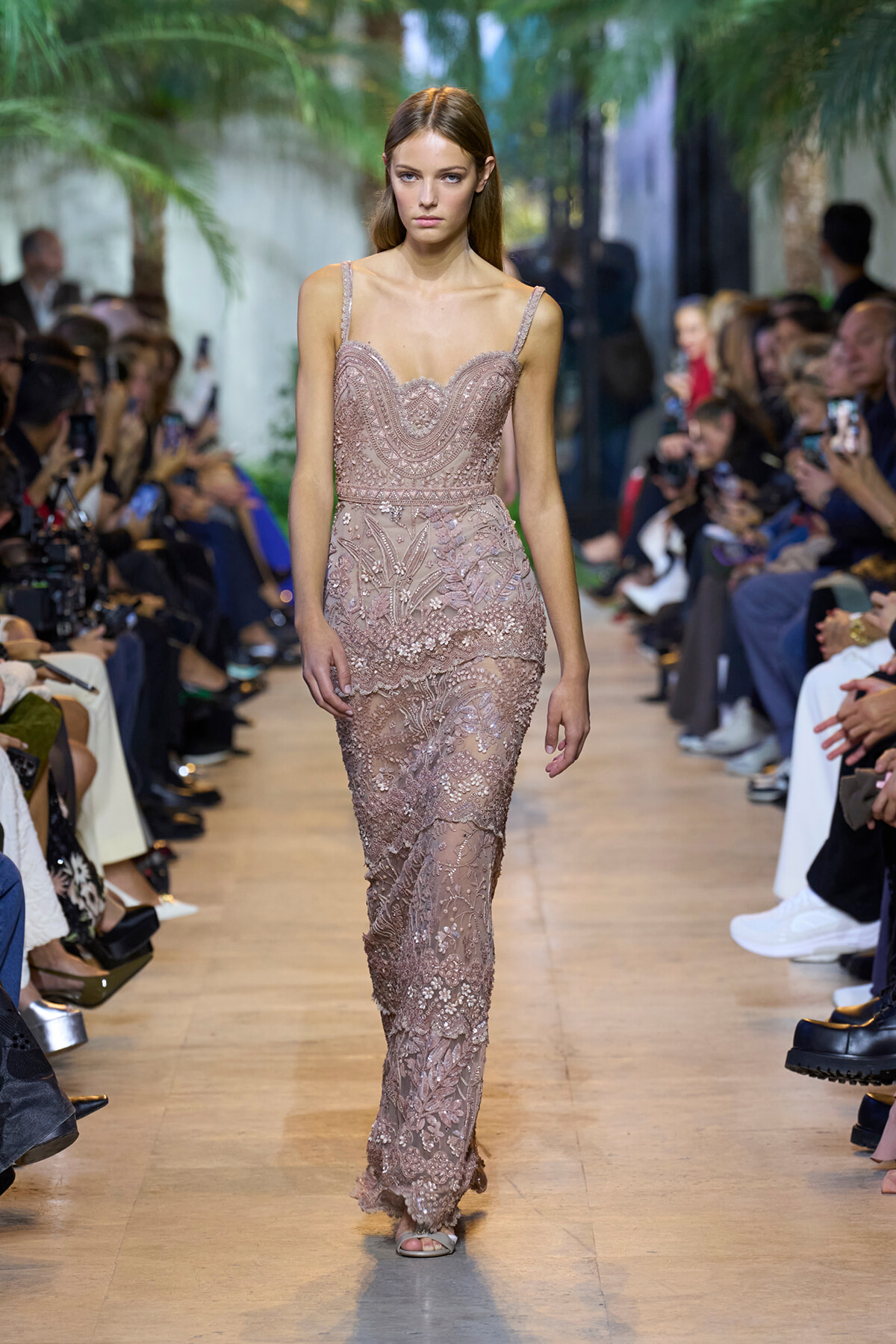 Model walks the runway in a blush pink beaded lace gown with intricate embellishments, audience on both sides.