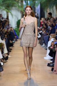 Model walks a fashion runway in a beige lace mini dress with a light gray shoulder bag, audience on both sides.