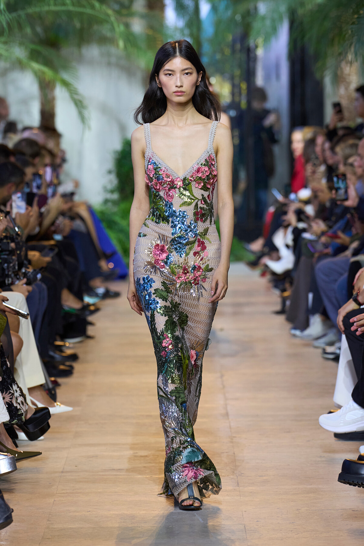 Model walks a runway in a sheer, floor-length gown with colorful floral embroidery and sequins, tropical backdrop visible on the sides.