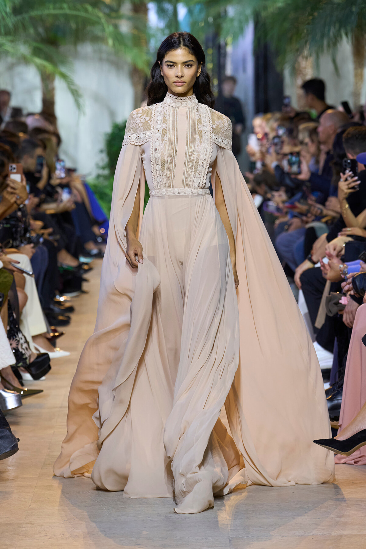 Model walking a runway in a floor-length beige gown with a lace bodice and dramatic cape sleeves.