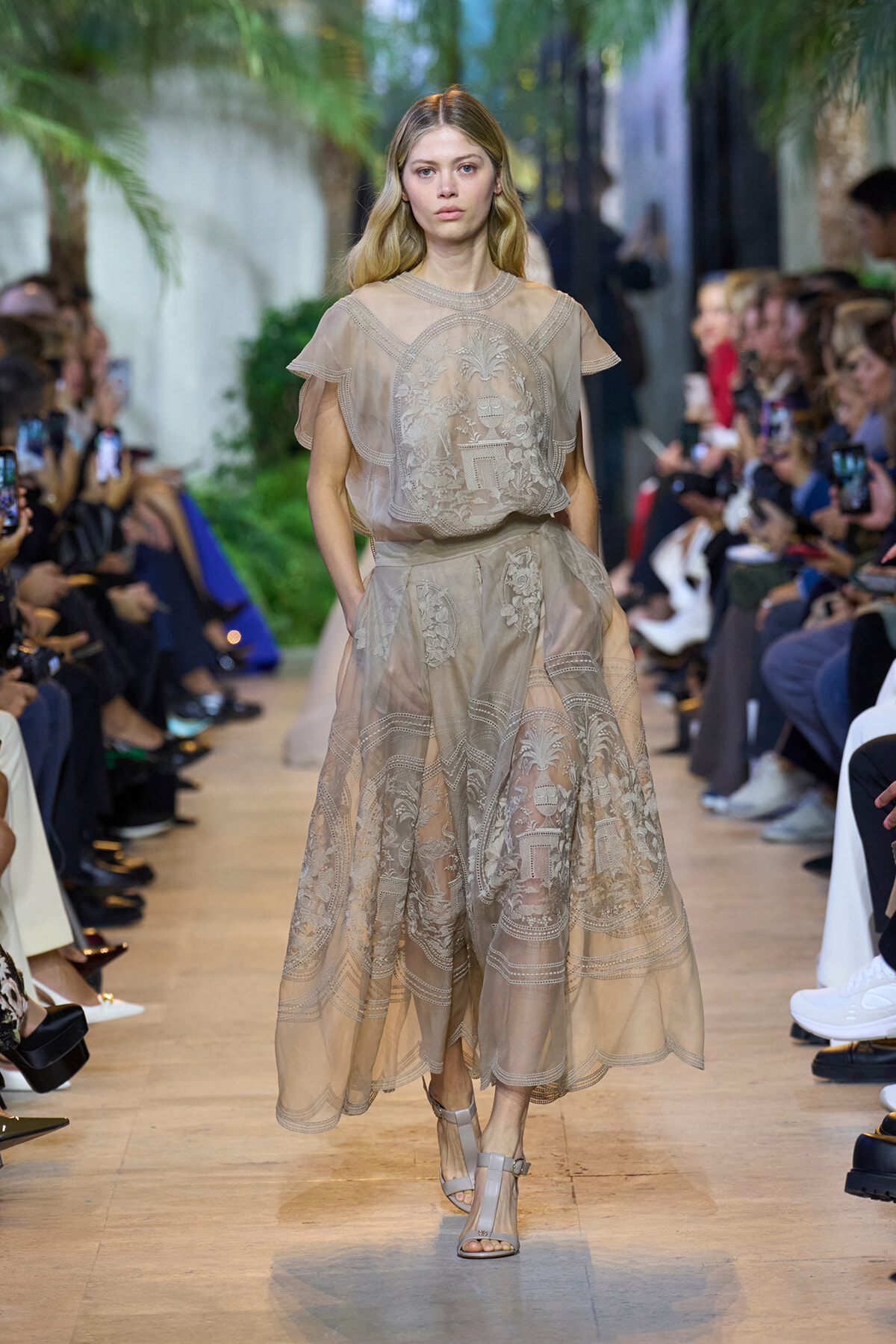 Model walks the runway in a beige sheer lace dress with floral embroidery, audience on both sides watching and filming.