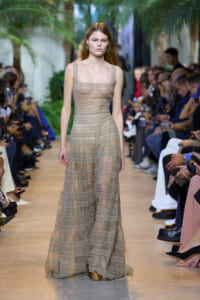 Model walking a runway in a long beige lace dress, crowd seated on both sides.