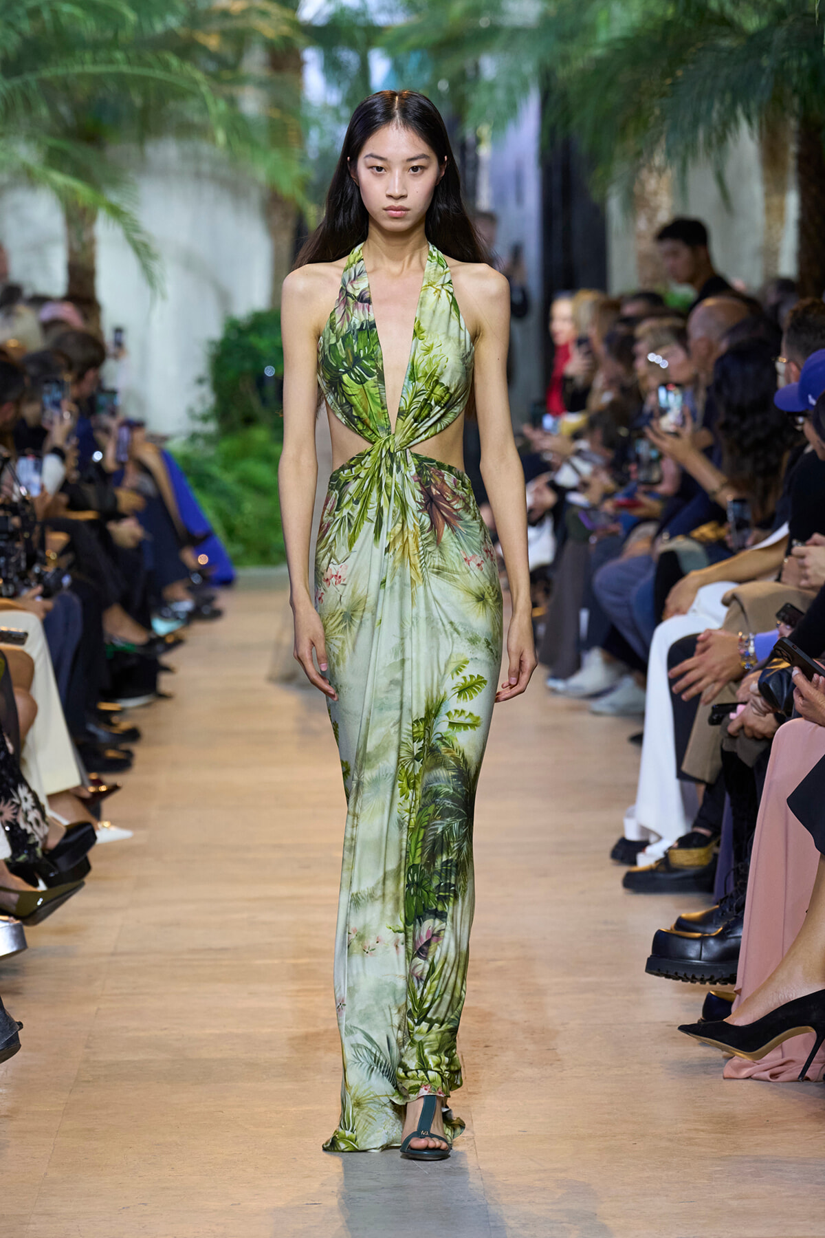 Model walks a tropical-green halter dress with a cutout waist down a fashion-show runway, audience on both sides.