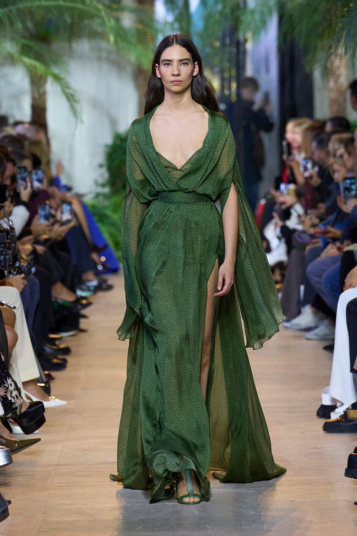 Model walks the runway in a green sheer draped gown with a deep V neckline and thigh-high slit, audience on both sides.] ,