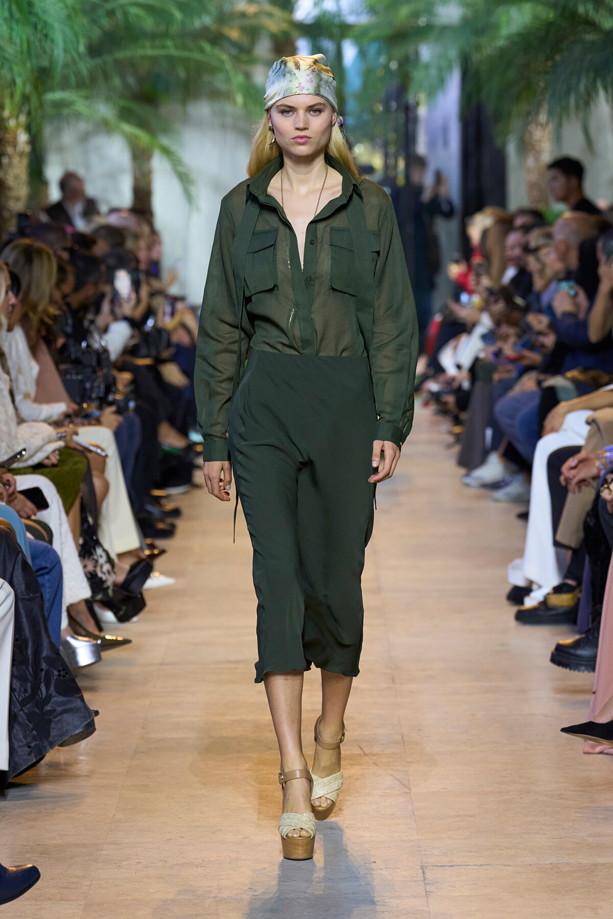 Model walks a fashion runway in a dark green utility jumpsuit with front pockets, paired with beige platform sandals and a pastel head scarf.