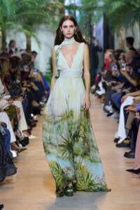 Model walks the runway in a pale green, tropical-print maxi dress with a scarf around the neck, audience on both sides.