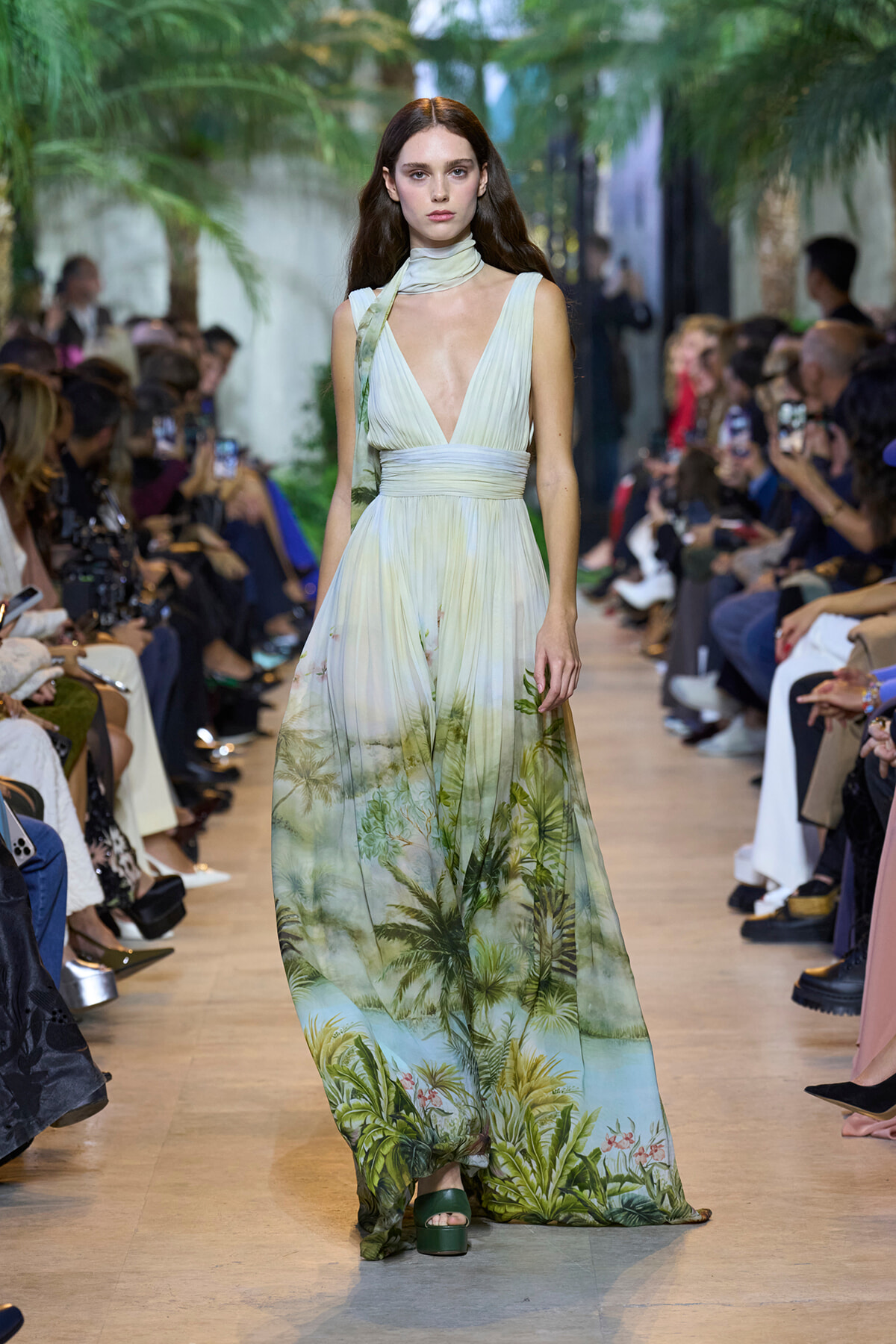 Model walks the runway in a pale green, tropical-print maxi dress with a scarf around the neck, audience on both sides.