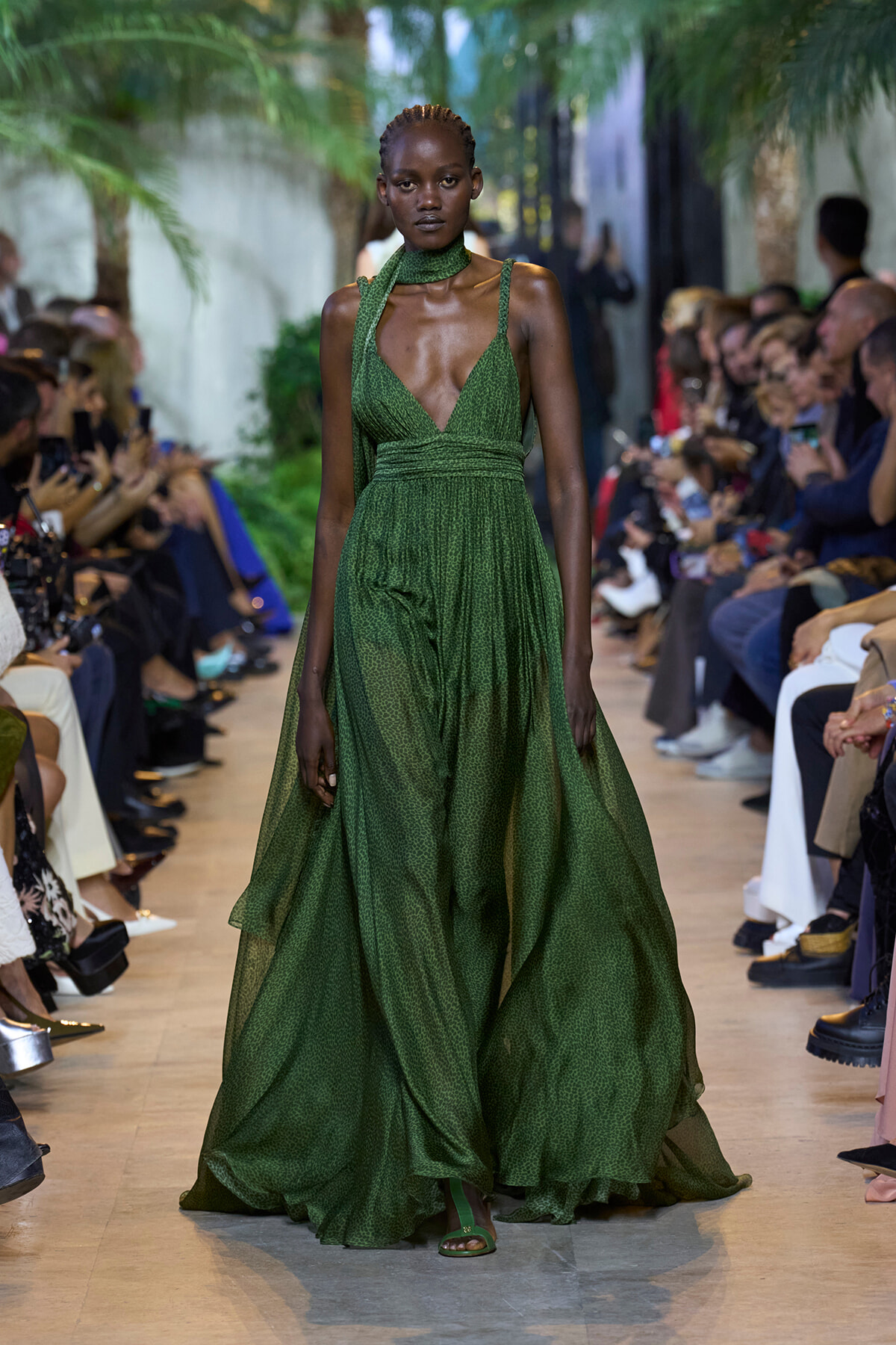 Model wearing an emerald-green halter gown with a plunging V-neck, walking a fashion runway.