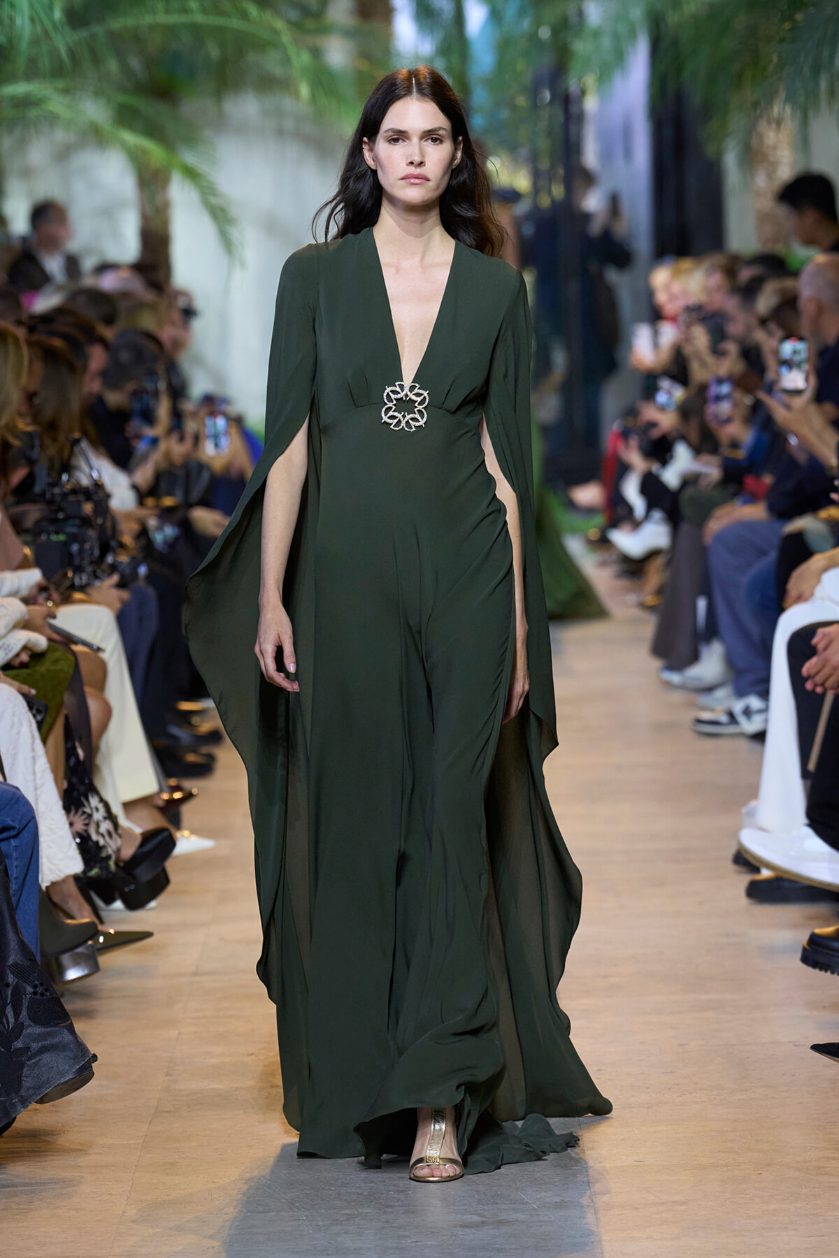 Model on a runway wearing a deep-green V-neck gown with cape sleeves and a silver floral waist brooch; floor-length and elegant.