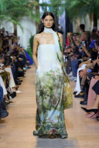 Model walks a runway in a sleeveless, light gradient maxi dress with a tropical palm print at the hem, on a garden-inspired catwalk.