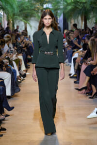Model walks a runway in a dark green tailored suit with a structured top, peplum waist, and wide belt.