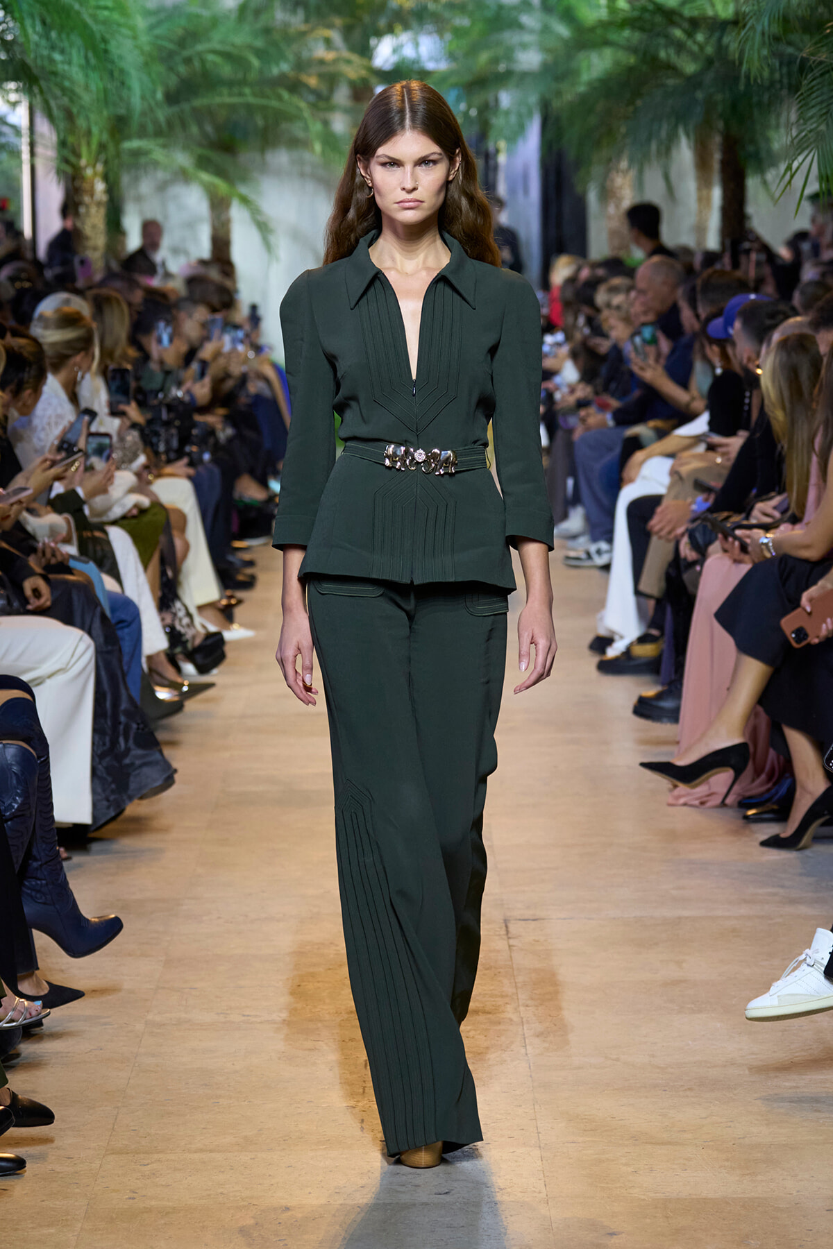 Model walks a runway in a dark green tailored suit with a structured top, peplum waist, and wide belt.