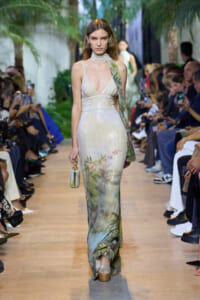 Model walks the runway in a shimmering, floor-length gown with a subtle floral print and a small metallic handbag in her left hand.