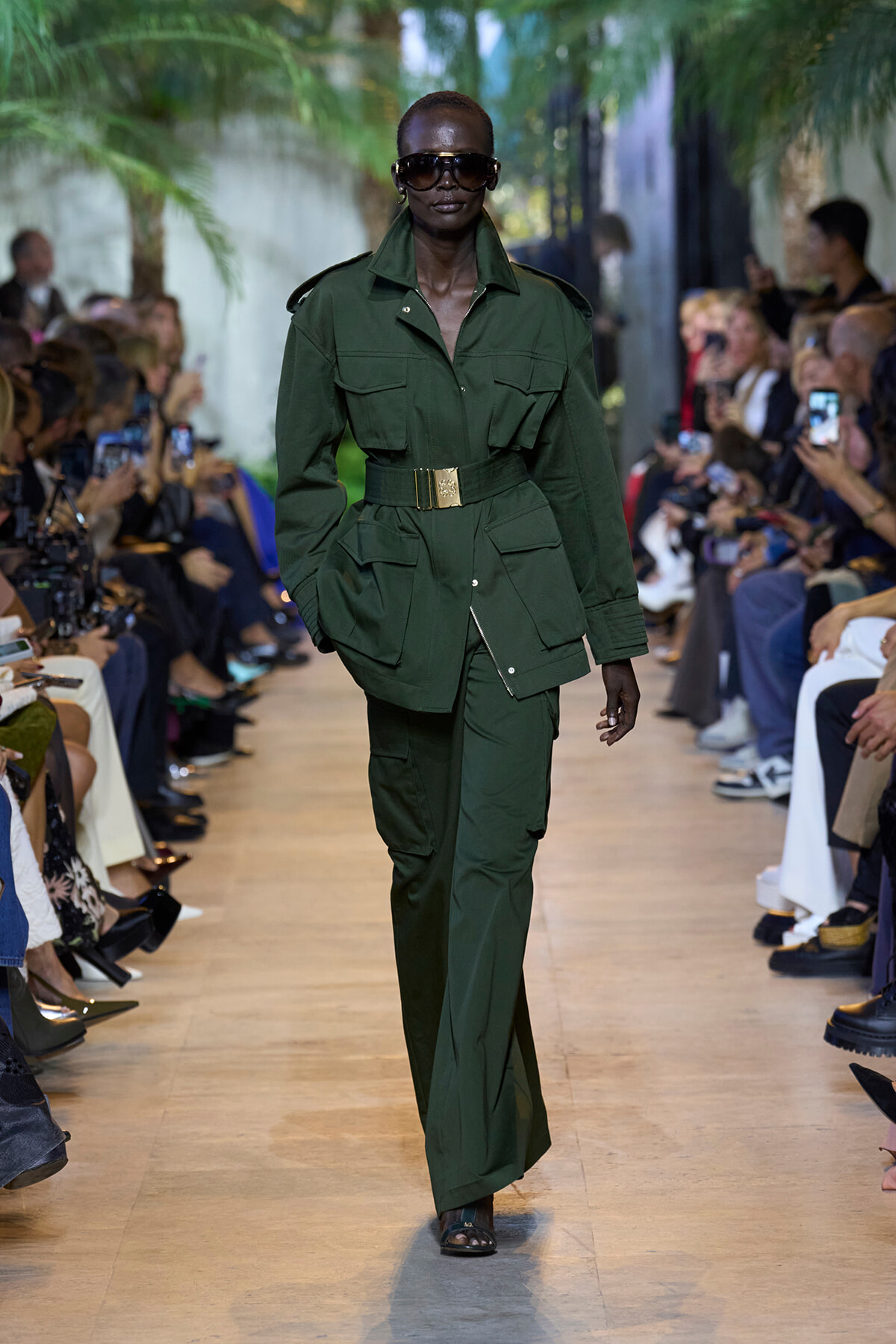 Model walks the runway in a forest-green utility suit with a gold belt and large pockets, wearing dark sunglasses.