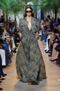 Model walking a fashion show in a full-length metallic snake-print dress with matching jacket and oversized sunglasses, hands in pockets.