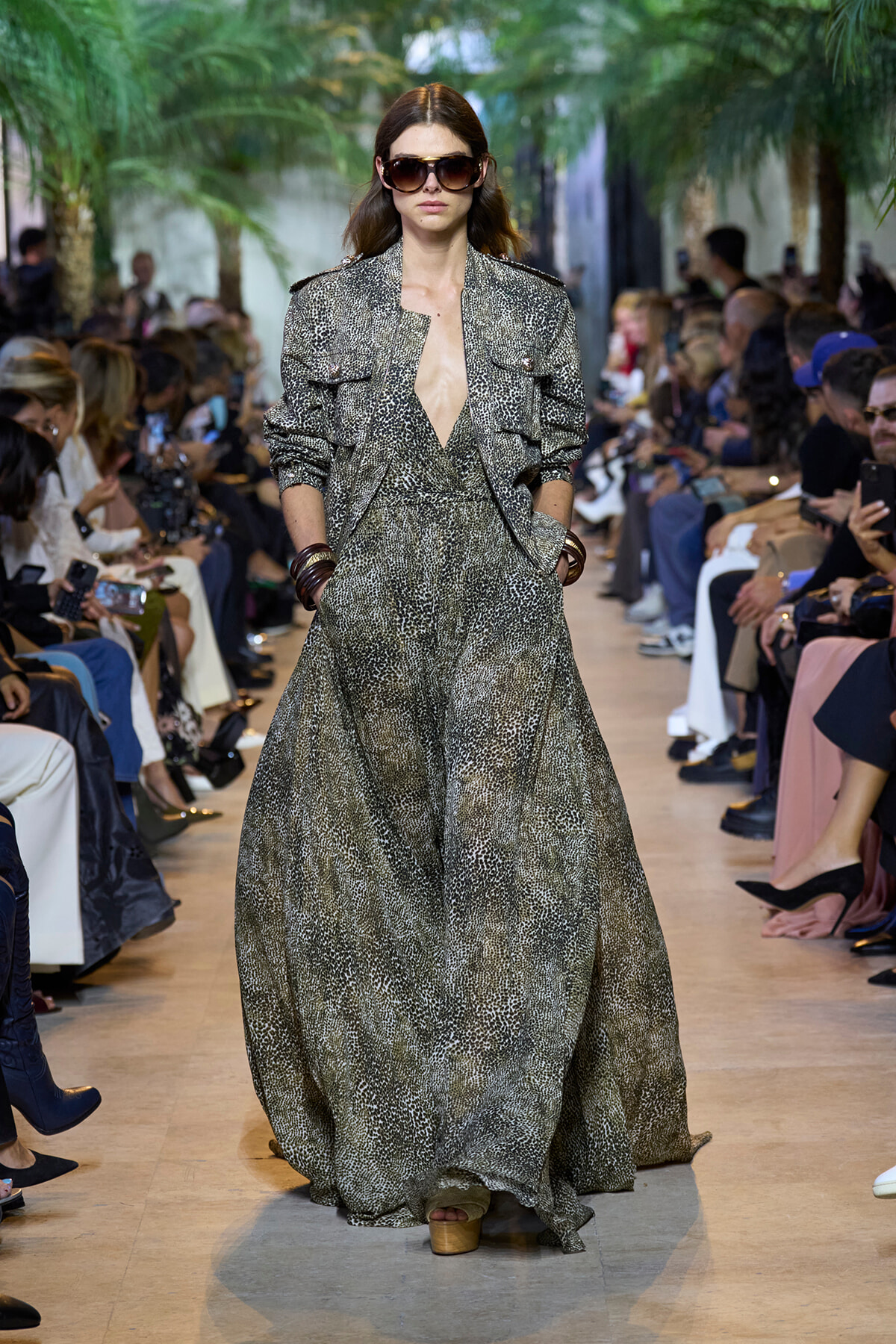 Model walking a fashion show in a full-length metallic snake-print dress with matching jacket and oversized sunglasses, hands in pockets.