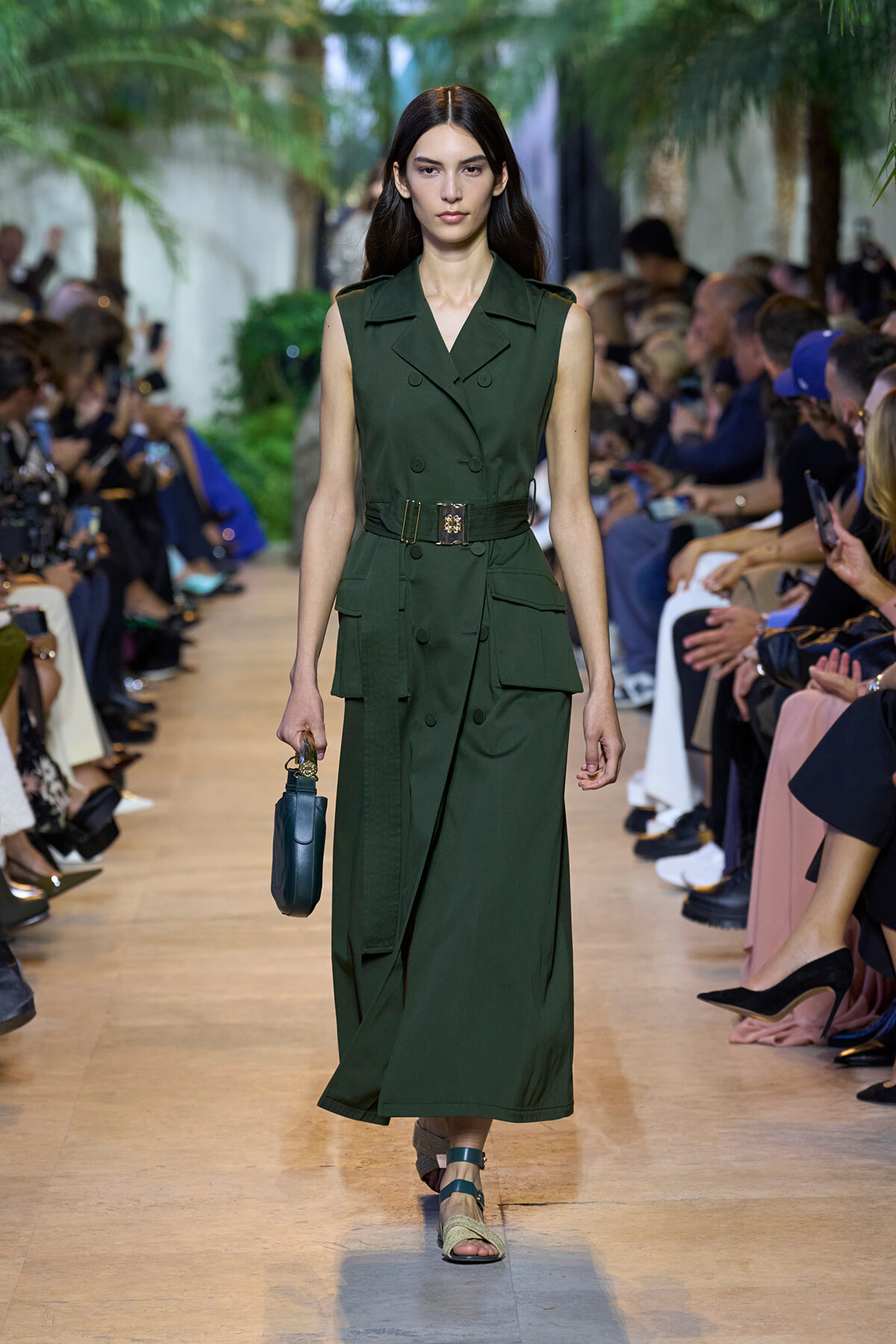 Model walking a green sleeveless trench dress with belt on a runway, holding a small blue handbag amid spectators.