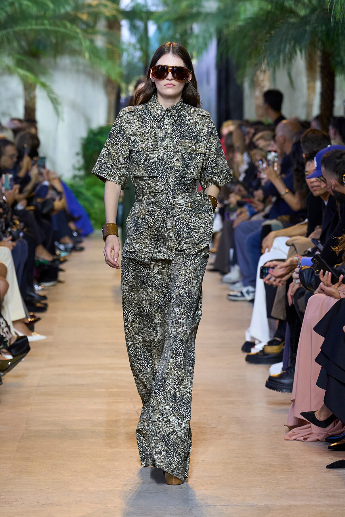 Model walking runway in a gray animal-print jumpsuit with wide legs and belt, wearing oversized sunglasses and brown cuffs, audience on both sides.