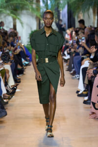 Model walks down a fashion runway in an olive green utility dress with a wide belt and sandals, audience on both sides take photos.