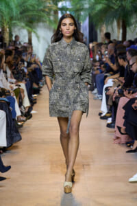 Model walks a runway wearing a metallic snakeskin-print shirt-dress with a belt, audience seated on both sides in a tropical-themed venue.