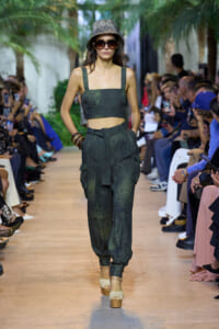 Model walking a runway in a dark green crop top and matching cargo pants, with a glitter bucket hat and oversized sunglasses.