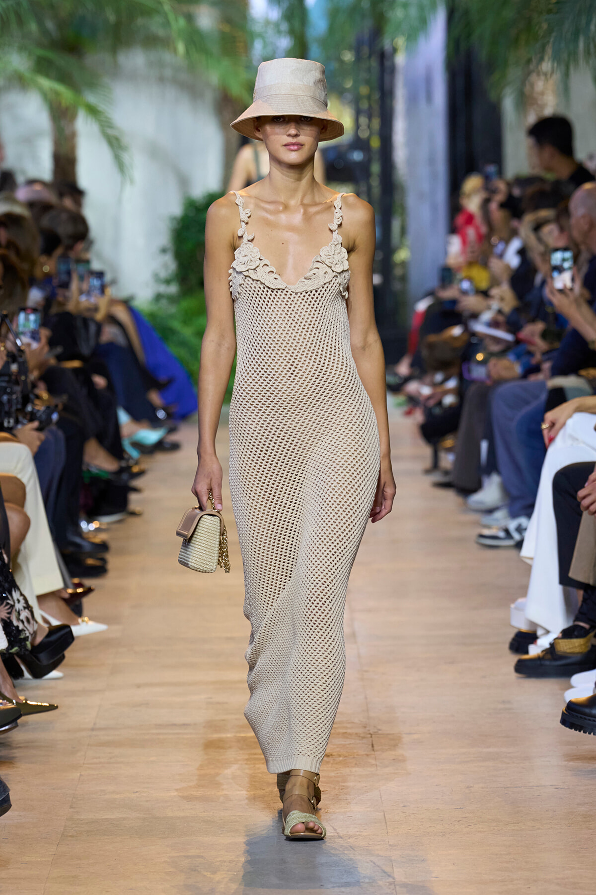 Model walks the runway in a beige crochet maxi dress with floral straps, paired with a wide-brim hat and woven handbag at a fashion show venue.