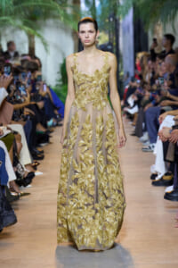 Model walks the runway in a beige gown with gold leaf embroidery and sheer panels, audience on both sides watching photographers.