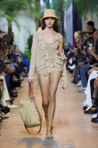 Model walks a fashion runway in a beige crochet ensemble with a matching hat, carrying a large woven tote with brown straps.