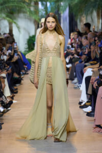 Model walks the runway in a beige crochet crop top and matching shorts with a flowing sheer skirt over them, carrying a crochet bag.