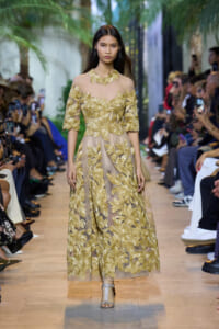 Model walks the runway in a sheer beige gown with gold leaf embroidery, audience filming nearby.
