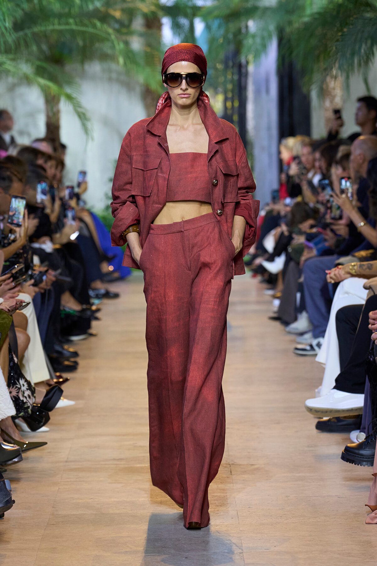 Model walking a fashion runway in a red coordinated suit with matching headscarf and sunglasses, audience on both sides.