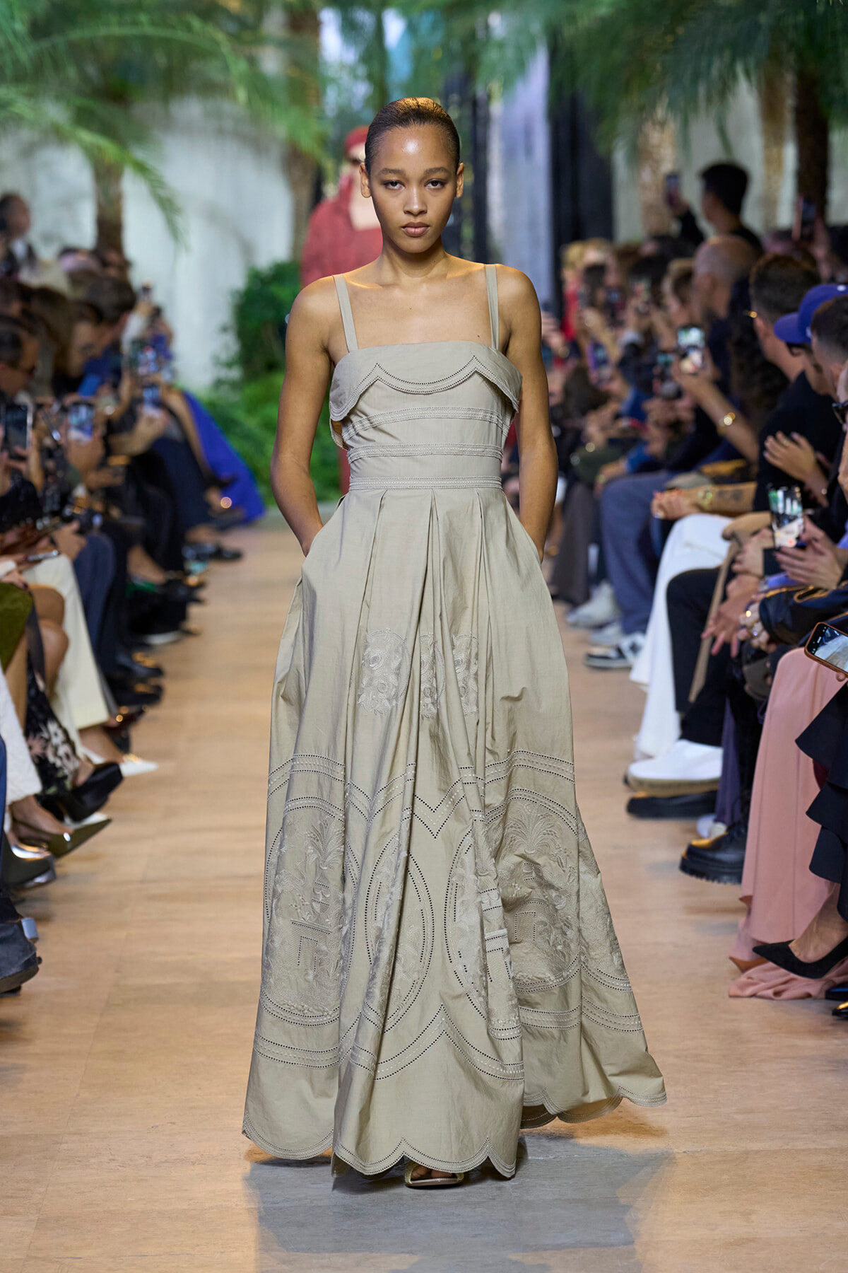Model on a fashion runway wearing a beige sleeveless maxi dress with embroidered panels; audience lines the catwalk and photographs.
